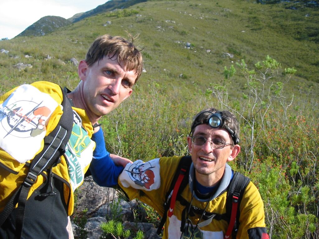 Two men standing in front of a mountain, looking at a camera